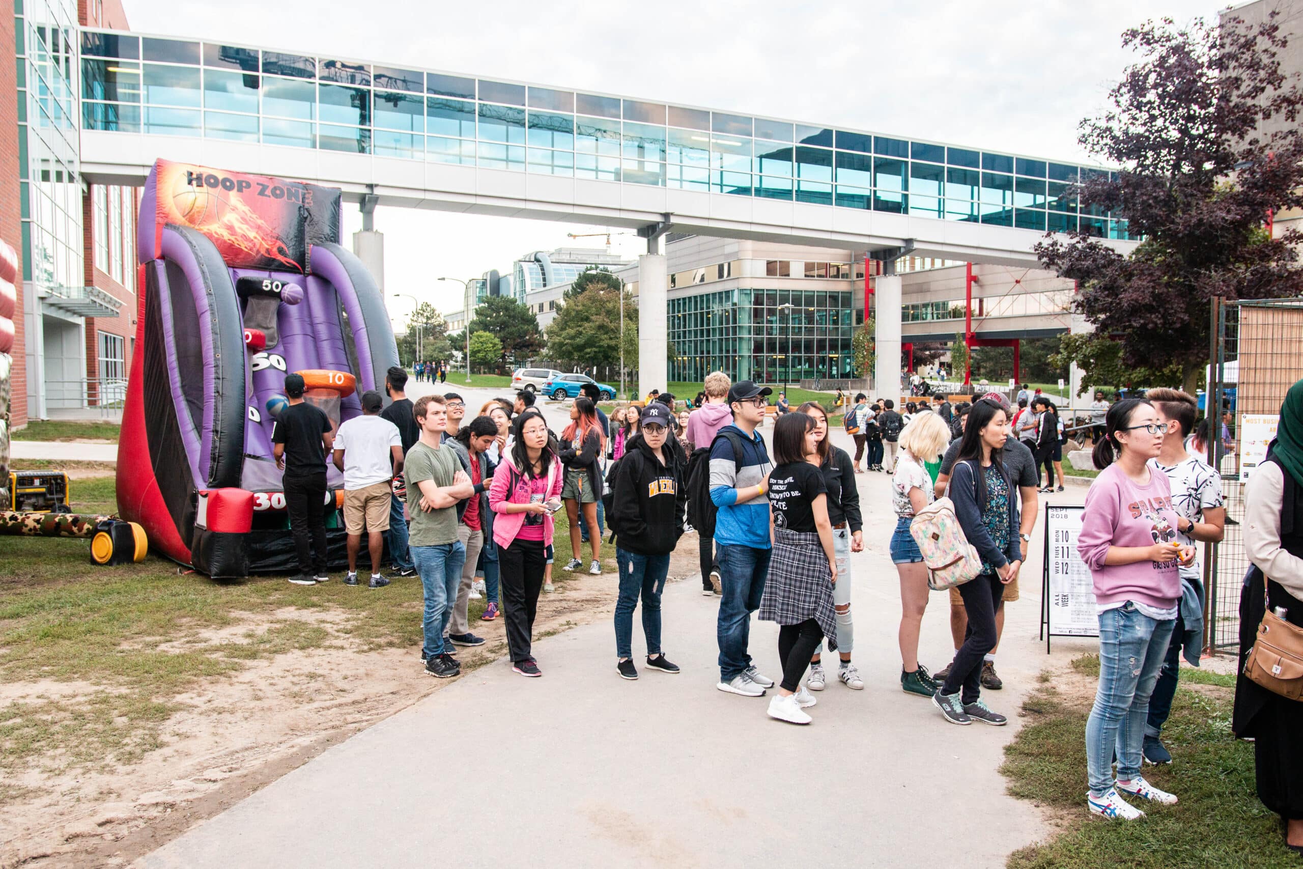 A diverse group of people stands in line outdoors on a campus, with some students chatting and others on their phones. Behind them is an inflatable basketball hoop game and modern campus buildings connected by a skybridge. Trees and greenery are visible around, typical of WUSA events' vibrant atmosphere.