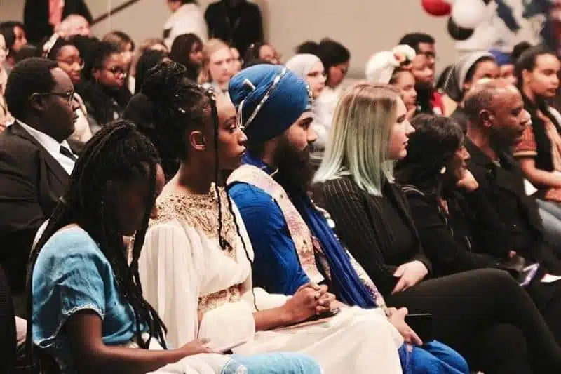 A diverse group of people are seated in an audience. Some attendees are in colorful traditional attire, while others wear more contemporary clothing. They appear to be focused on a presentation or speech. The setting suggests a formal event or conference.