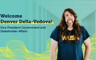 A person with long hair and a mustache, wearing a blue T-shirt, stands in front of a teal background with abstract yellow wave designs. The text reads, "Welcome Denver Della-Vedova! WUSA Vice President Government and Stakeholder Affairs.