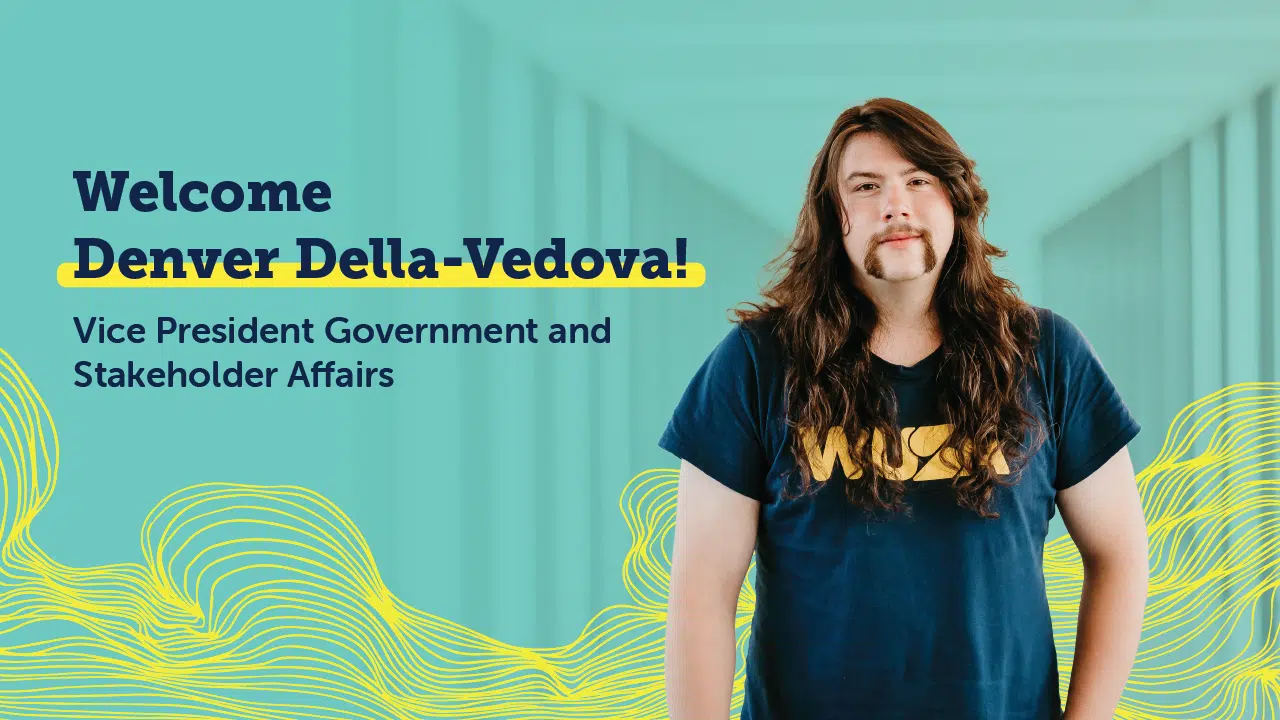 A person with long hair and a mustache, wearing a blue T-shirt, stands in front of a teal background with abstract yellow wave designs. The text reads, "Welcome Denver Della-Vedova! WUSA Vice President Government and Stakeholder Affairs.