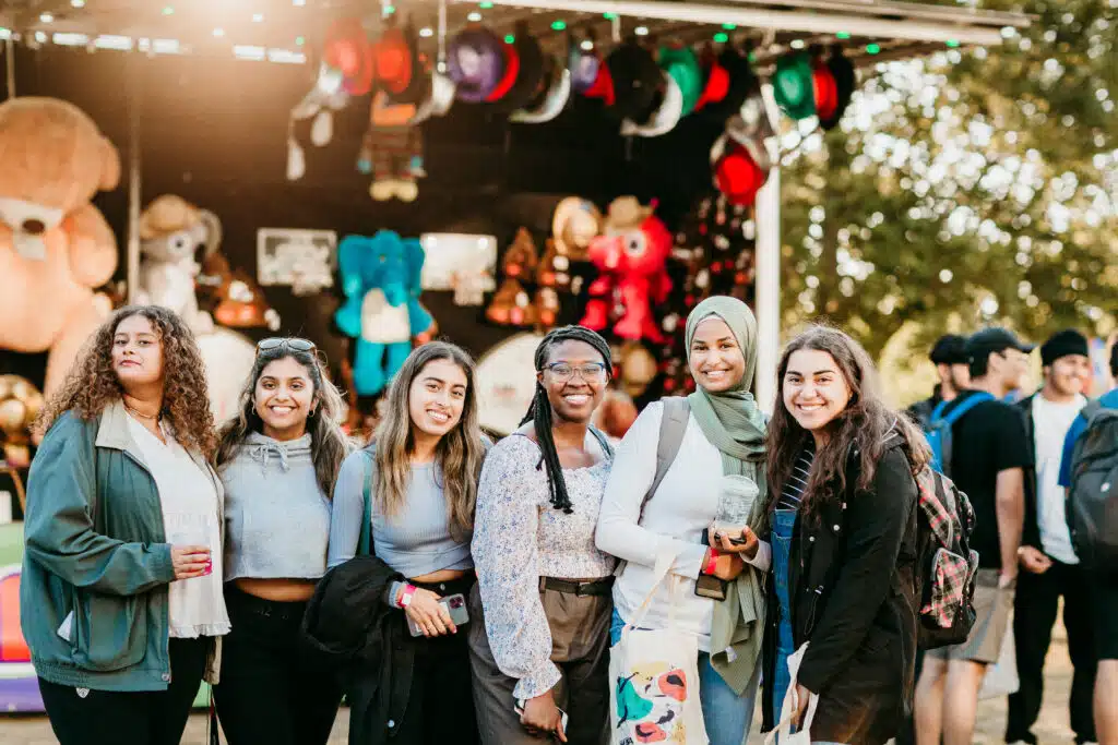 A group of six diverse women standing and posing for a photo at what appears to be an outdoor carnival or fair. They are smiling and dressed casually, with stuffed animals and colorful hats displayed in the background. Trees and other people are visible in the distance.