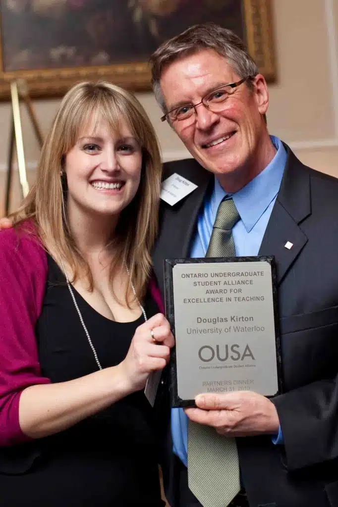 A woman and a man are smiling and holding an award plaque together. The plaque reads, "Ontario Undergraduate Student Alliance Award for Excellence in Teaching, Douglas Kirton, University of Waterloo." They are both dressed formally.