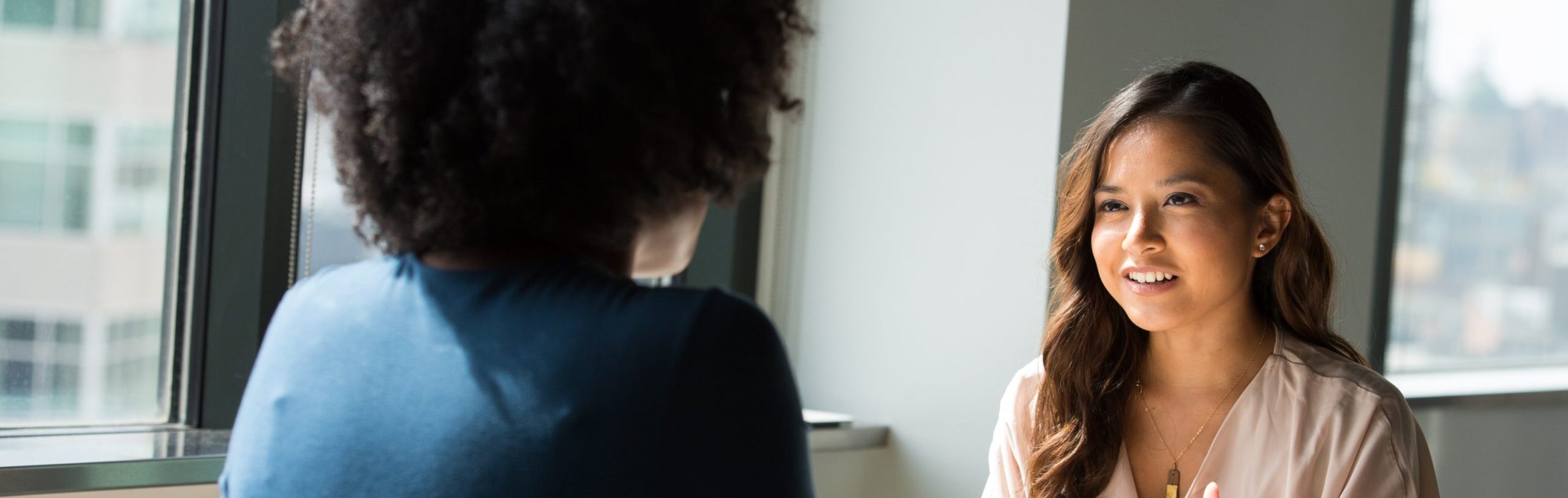 Two women sit at a table facing each other, engaged in a consultation. The woman on the left, with long brown hair and a light-colored blouse, is speaking and gesturing with her hands. The woman on the right, who has curly hair and is wearing a dark-colored top, listens intently.