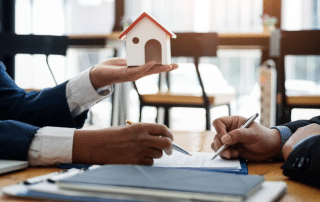 Two people wearing business suits are engaged in a discussion at a table. One person is holding a small model of a house, while the other is writing on a document. The background shows a window with natural light.