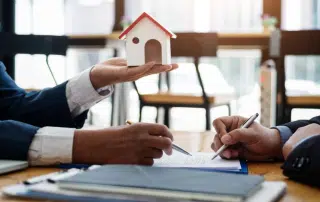 Two people wearing business suits are engaged in a discussion at a table. One person is holding a small model of a house, while the other is writing on a document. The background shows a window with natural light.