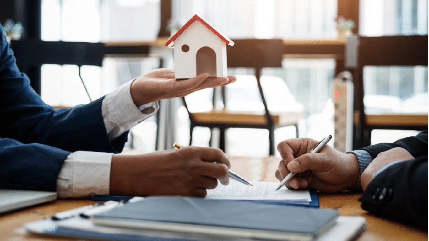 Two people wearing business suits are engaged in a discussion at a table. One person is holding a small model of a house, while the other is writing on a document. The background shows a window with natural light.
