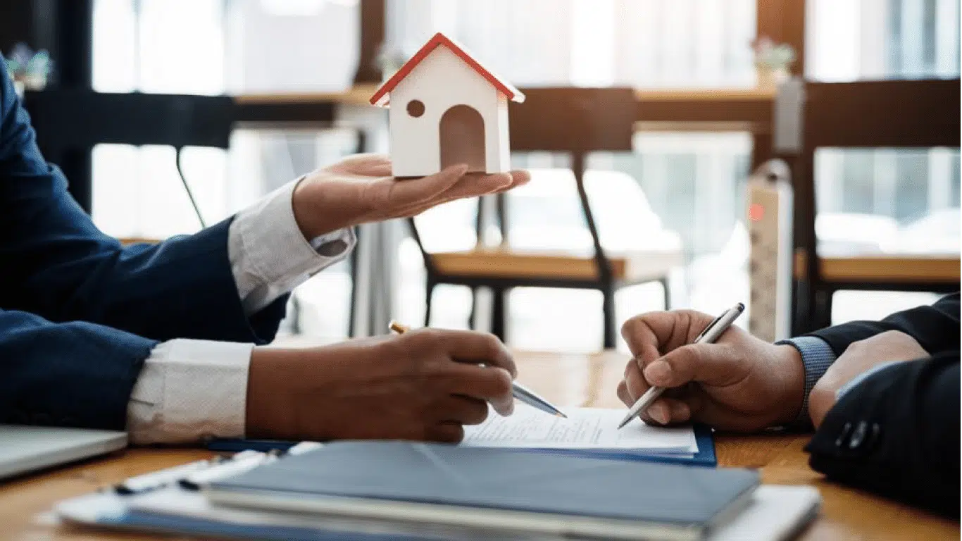 Two people wearing business suits are engaged in a discussion at a table. One person is holding a small model of a house, while the other is writing on a document. The background shows a window with natural light.
