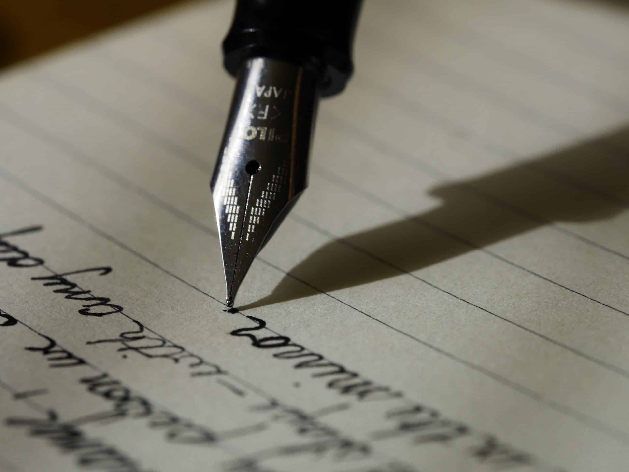 Close-up of a fountain pen writing cursive text on lined paper. The pen nib is metallic, and the ink is dark, making a shadow as it writes. The handwritten text appears in an elegant and flowing script, though the specific words are not fully readable.
