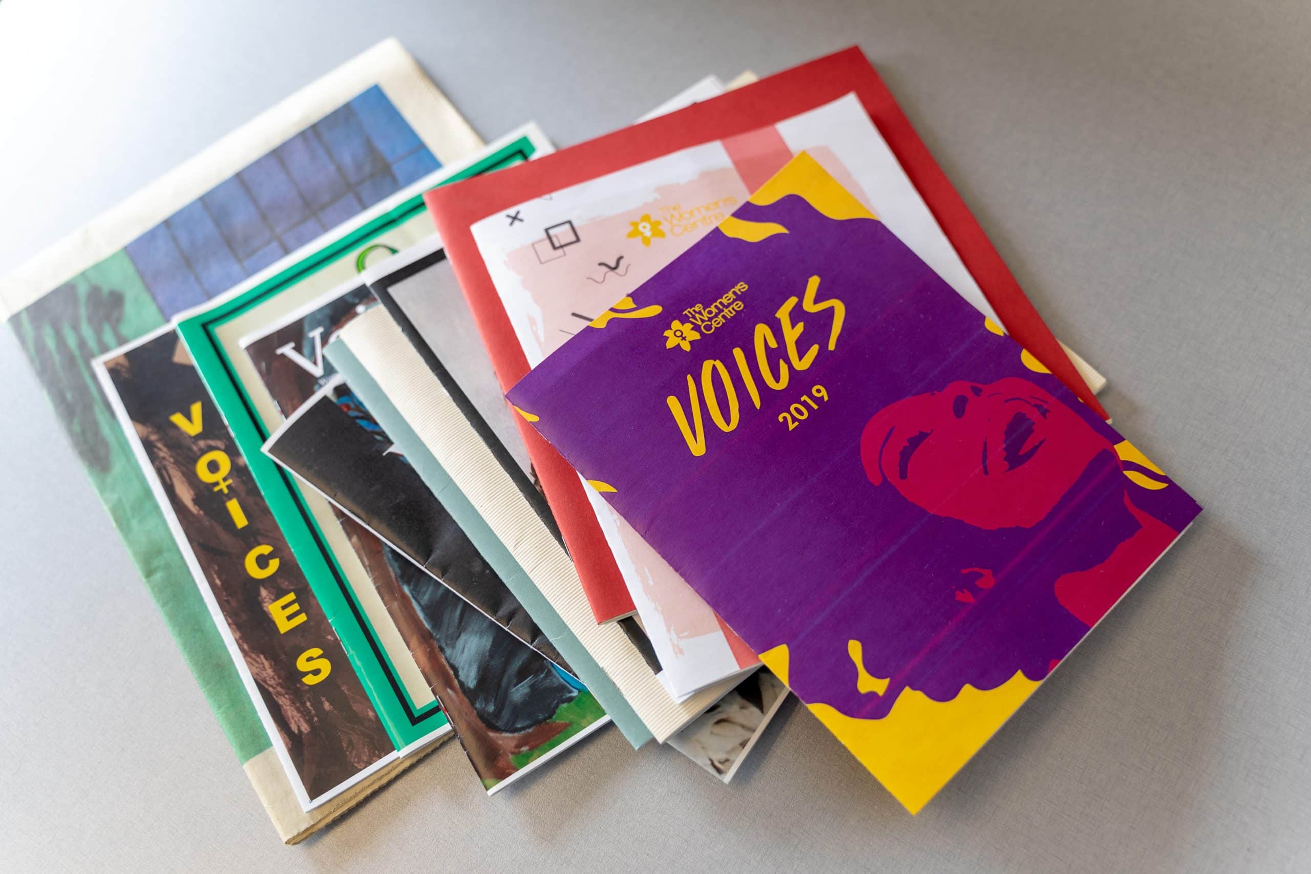 A stack of colorful, assorted booklets and pamphlets is spread on a flat surface. The top booklet prominently features an abstract design of a person's face and the word "VOICES 2019" in bold yellow letters on a purple background.