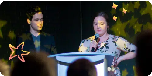 A man and a woman are standing on a stage. The woman is speaking into a microphone while the man looks on. The background features a star-themed projection and illustrative star graphics.