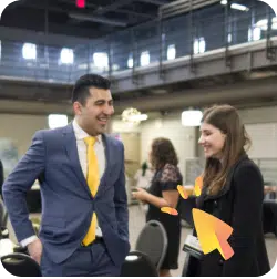 Two people are engaging in a lively conversation indoors. The man on the left is dressed in a blue suit with a yellow tie, and the woman on the right is wearing a black outfit. Both appear to be smiling. The background features other individuals and tables set up for an event.