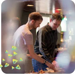 Two people standing side by side at a catered event, serving themselves food from a buffet table. One person is wearing a striped T-shirt and the other is in a dark shirt. The scene is warm and friendly, with soft lighting and a blurred foreground framing the image.