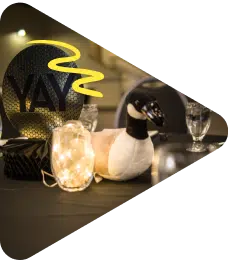 A decorated banquet table with a plush toy goose, a lit glass jar, and a black balloon with the word "YAY" in bold yellow letters. The items are arranged on a dark tablecloth with chairs in the background.