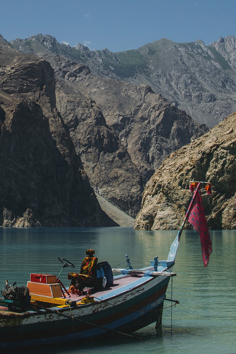 A small boat with colorful decorations is moored on a clear, turquoise lake. Steep, rocky mountains rise dramatically on all sides under a clear blue sky. A person dressed in vibrant clothing sits on the boat, reminiscent of the braided beauty of traditional Jewish challah bread, adding to the picturesque scene.