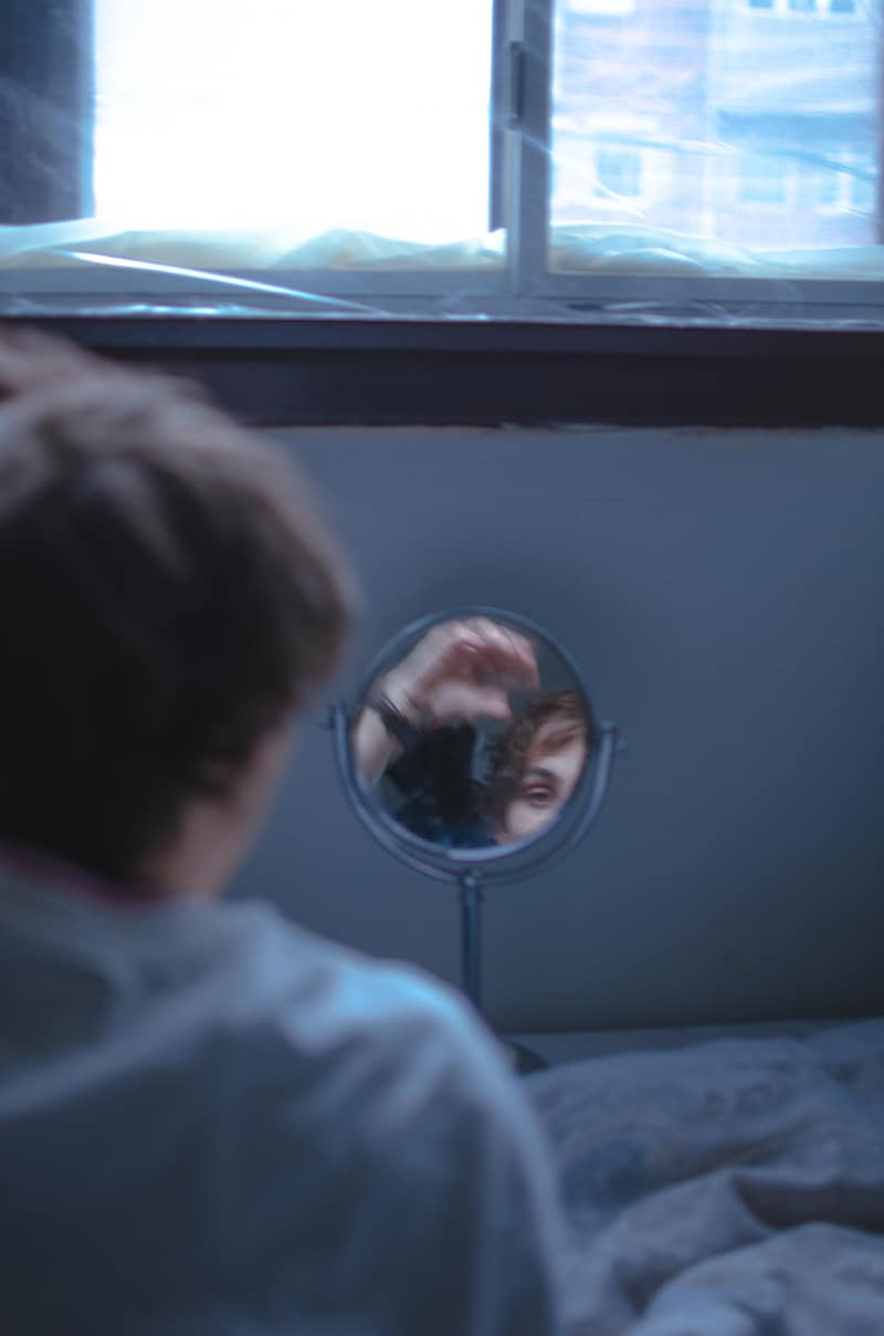 A person with short hair is sitting in front of a small round mirror placed on a table, resembling the meticulous poise of a Million Dollar Man. The mirror shows their reflection as they lift a hand to adjust their hair. The background includes a window with frosted glass, and the scene appears softly lit and blurry.