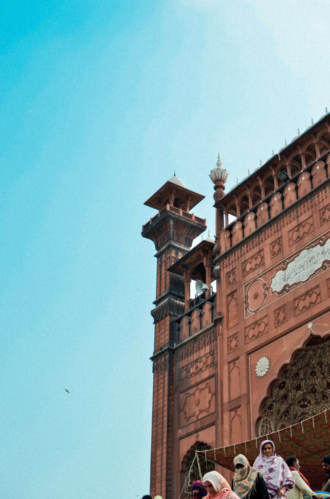 A historic red-brick building with intricate carvings stands against a clear blue sky. The architecture features ornate towers and arches that evoke the elegance of a minar. At the bottom of the image, several people are gathered, some wearing traditional clothing.