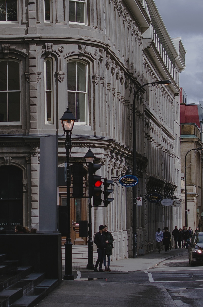 A city street in Montreal featuring historic buildings with ornate detailing. A few people are walking on the sidewalk, and two individuals stand at a crosswalk waiting for the traffic light to change. The light is currently red. Street lamps and store signs are visible, giving mtl its charming vibe.