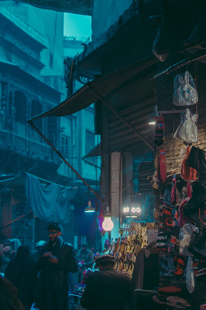 A dimly lit street market scene at the Nurian Bazaar, with various items, such as hats and shoes, hanging from a stall in the foreground. Shoppers and vendors are visible in the background. The architecture has an old-fashioned style with intricate details. The atmosphere is bustling yet moody.