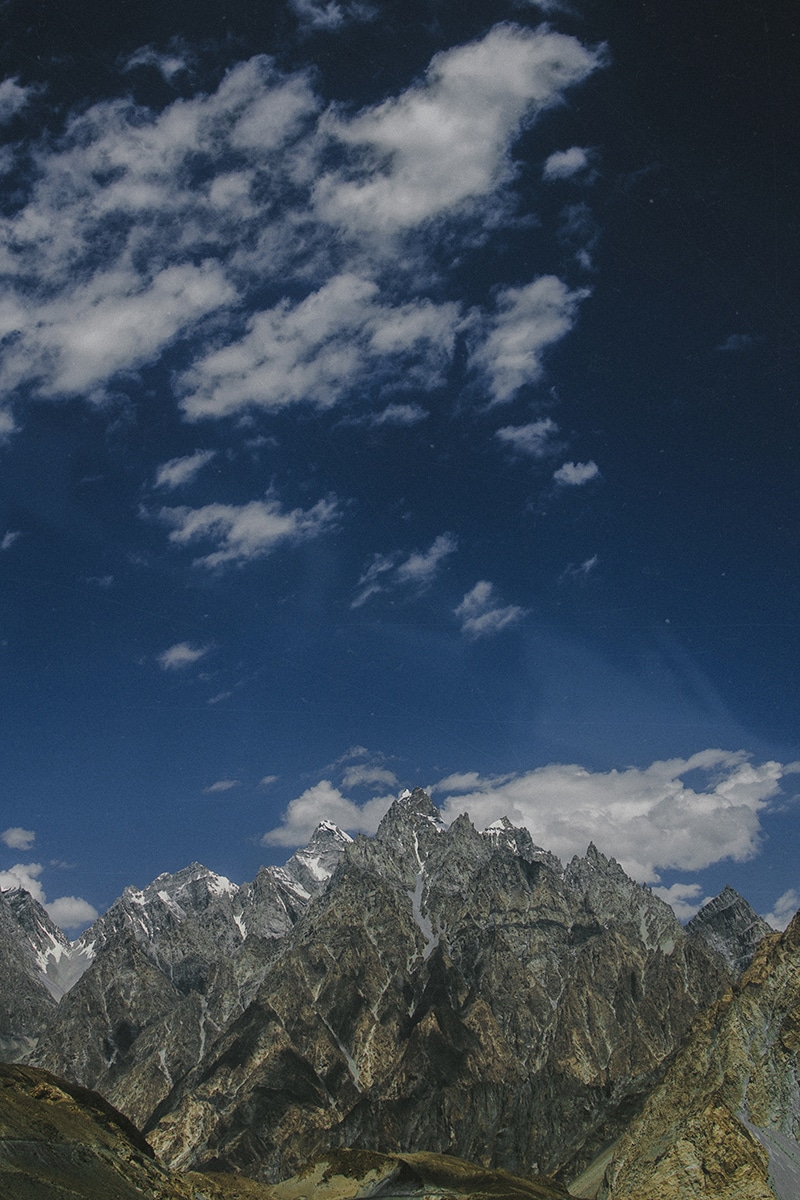 A dramatic mountain range with rugged, jagged peaks stretching under a vibrant, deep blue sky scattered with wispy white clouds. The stark contrast between the rocky Passu mountains and the clear sky creates a striking and awe-inspiring natural landscape.