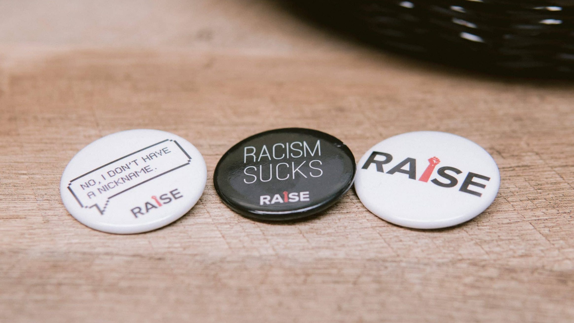 Three circular buttons on a wooden surface. The left button is white with text: "NO, I DON'T HAVE A NICKNAME." The middle button is black with white text: "RACISM SUCKS." The right button is white with "RAISE" written in black and red.