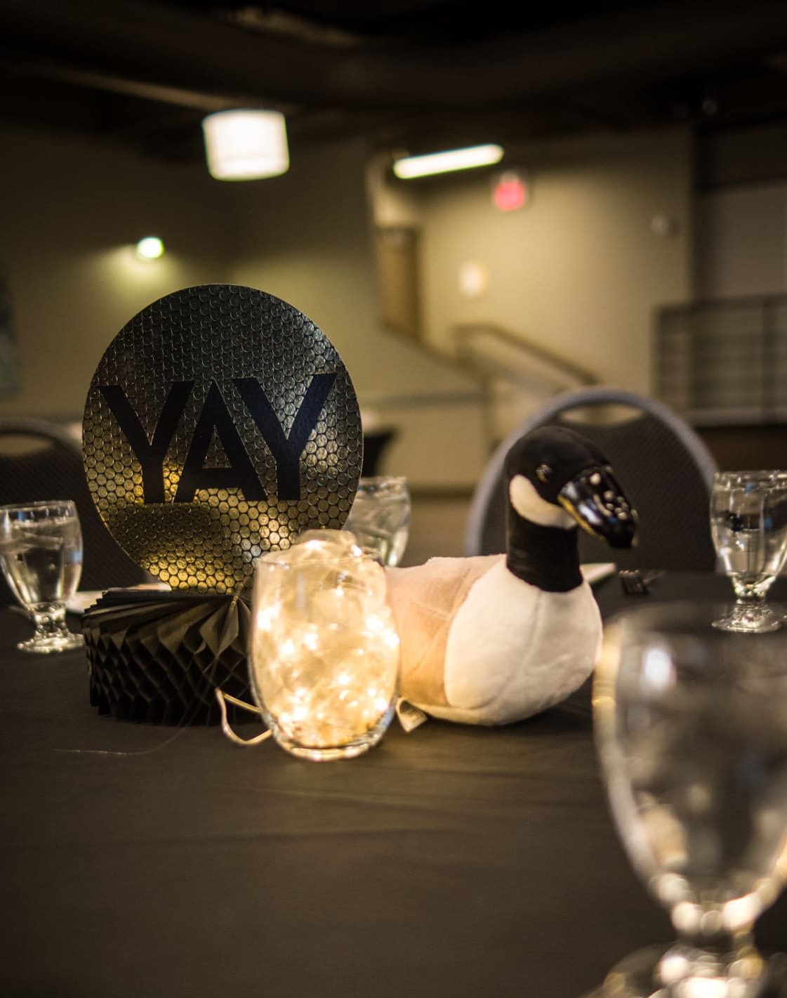 A table centerpiece featuring a stuffed Canada goose, a round black sign with "YAY" written on it, and a jar filled with string lights. The tabletop also has glasses of water and a black honeycomb paper decoration, all set in a dimly lit room.