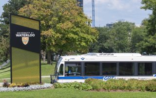 A blue and white city bus passes by a large University of Waterloo sign on a green lawn. The sign features the university's crest and name. Trees and buildings are visible in the background under a partly cloudy sky.