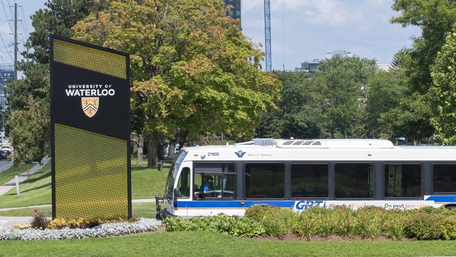 A blue and white city bus passes by a large University of Waterloo sign on a green lawn. The sign features the university's crest and name. Trees and buildings are visible in the background under a partly cloudy sky.