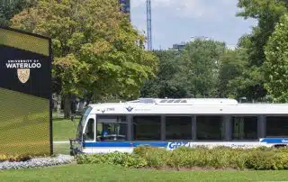 A white and blue city bus is parked near a sign that reads "University of Waterloo." The sign is surrounded by greenery and flowers. Buildings and trees are visible in the background, along with a clear sky.