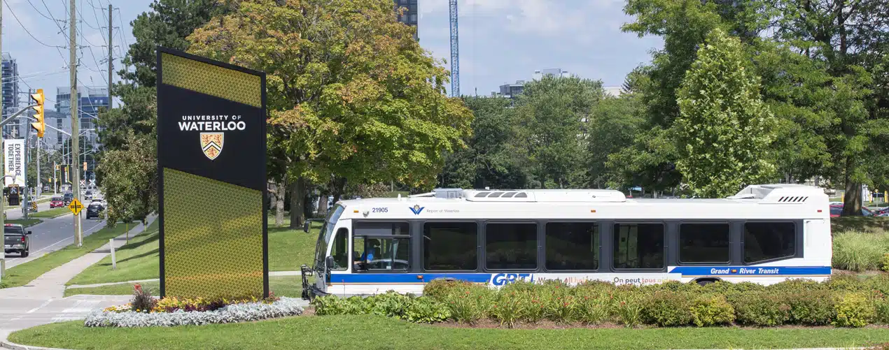 A white and blue city bus is parked near a sign that reads “University of Waterloo.” The sign is surrounded by greenery and flowers. Buildings and trees are visible in the background, along with a clear sky. A white and blue city bus is parked near a sign that reads "University of Waterloo." The sign is surrounded by greenery and flowers. Buildings and trees are visible in the background, along with a clear sky.