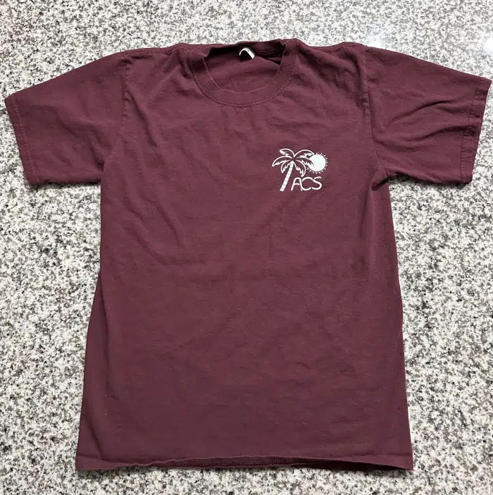 An Association of Caribbean Students (ACS) T-shirt in maroon is laid flat on a speckled stone surface. The shirt, designed for Caribbean Students, features a small graphic on the upper left chest area with a palm tree, a sun, and the letters "ACS" underneath the tree.