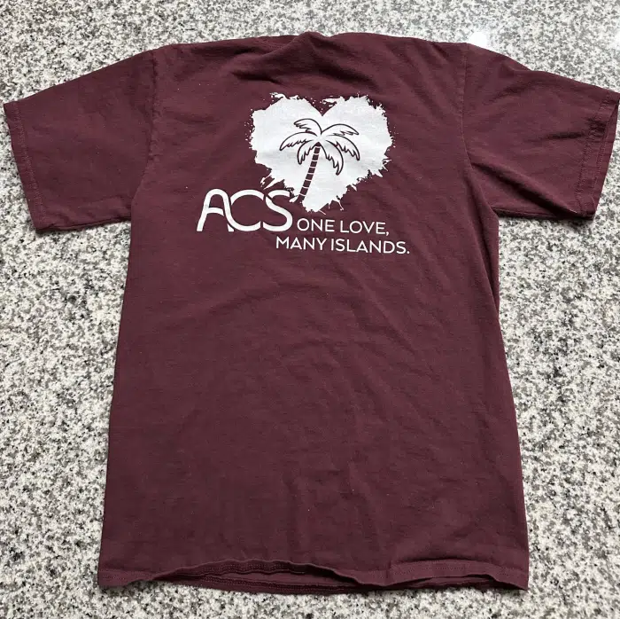 An Association of Caribbean Students (ACS) T-shirt in maroon lies on a gray speckled surface. The shirt features a white graphic of a palm tree within a heart-shaped design and the text "ACS One Love, Many Islands," celebrating the unity of Caribbean students through the association.