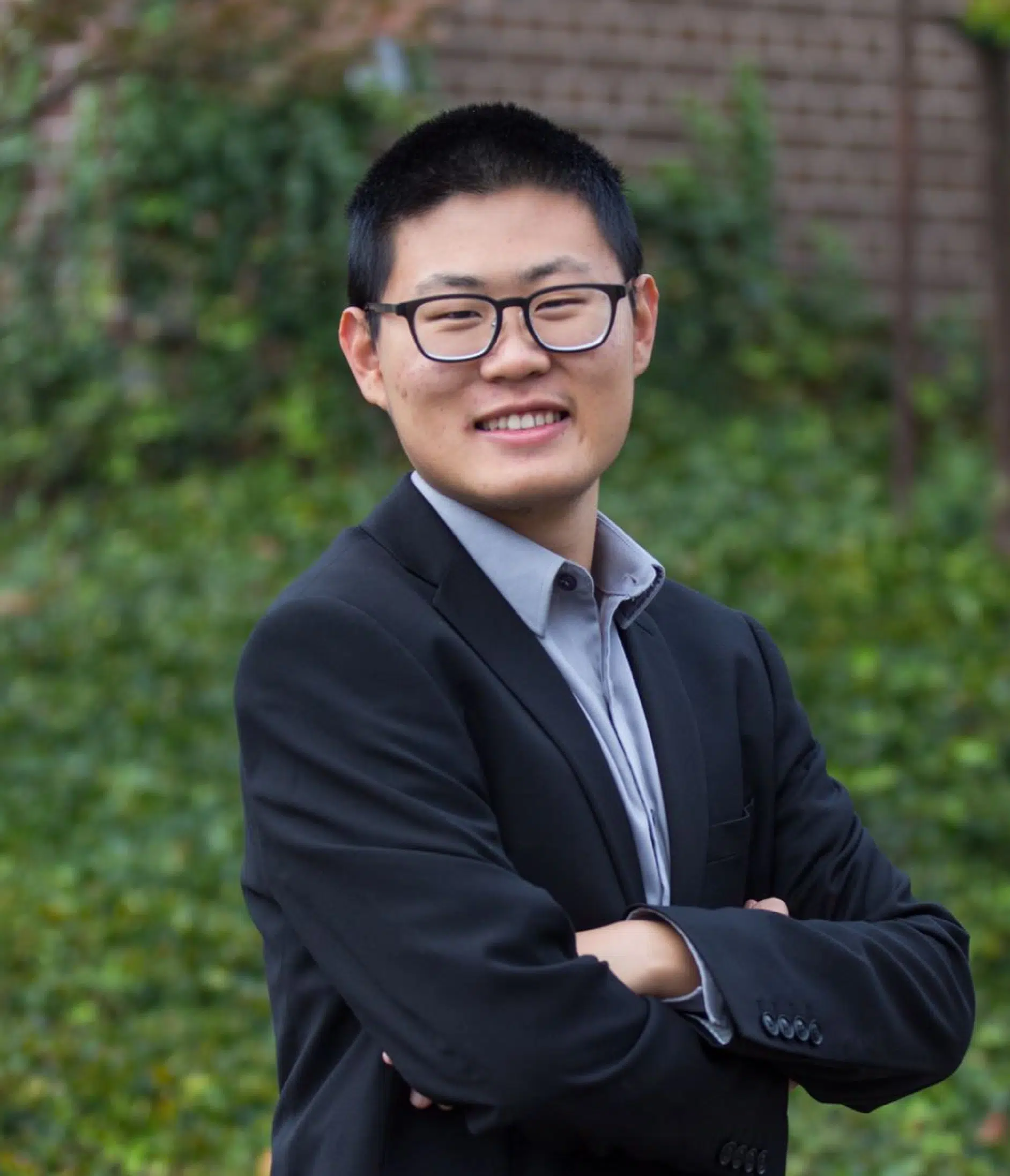 A person with short black hair and glasses is smiling and standing outdoors with arms crossed, wearing a black blazer over a light blue shirt. The background features greenery and a brick wall.