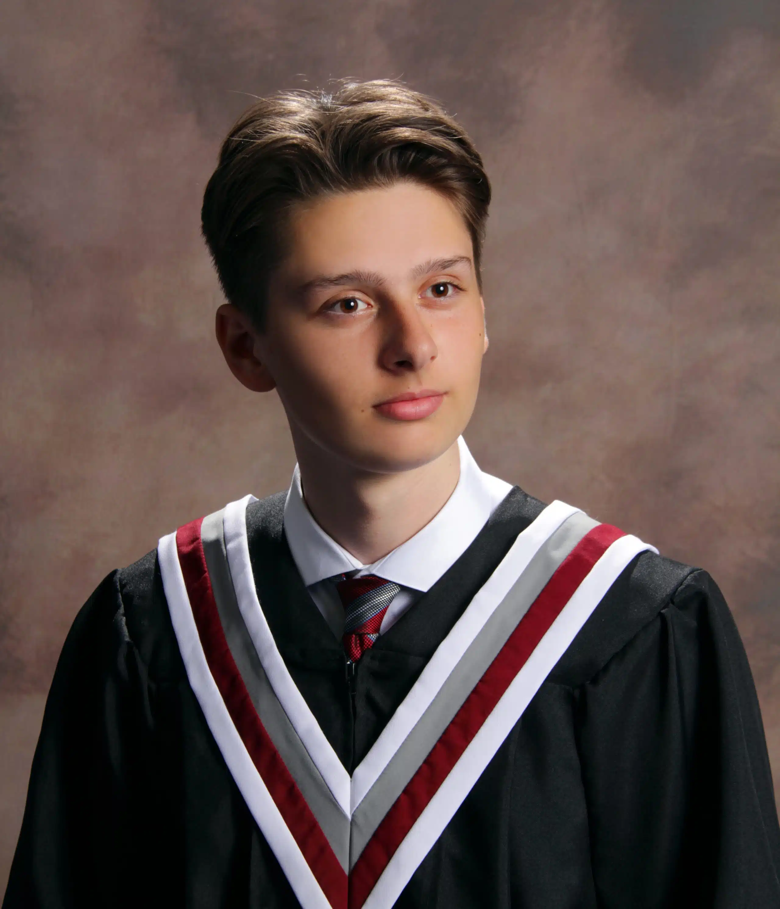 A young person with short brown hair is wearing a black graduation gown with white and red trim, a white collared shirt, and a red and grey striped tie. They are looking slightly to the side with a neutral expression. The background is a muted brown and grey.