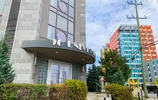 A modern building entrance with the address "96 University" displayed above the door. The entrance features glass blocks and a circular accent over the double doors. Surrounding the building are bushes and other buildings, with colorful, uniquely designed structures visible. The location is notable for hosting significant events such as the August Board Meeting.