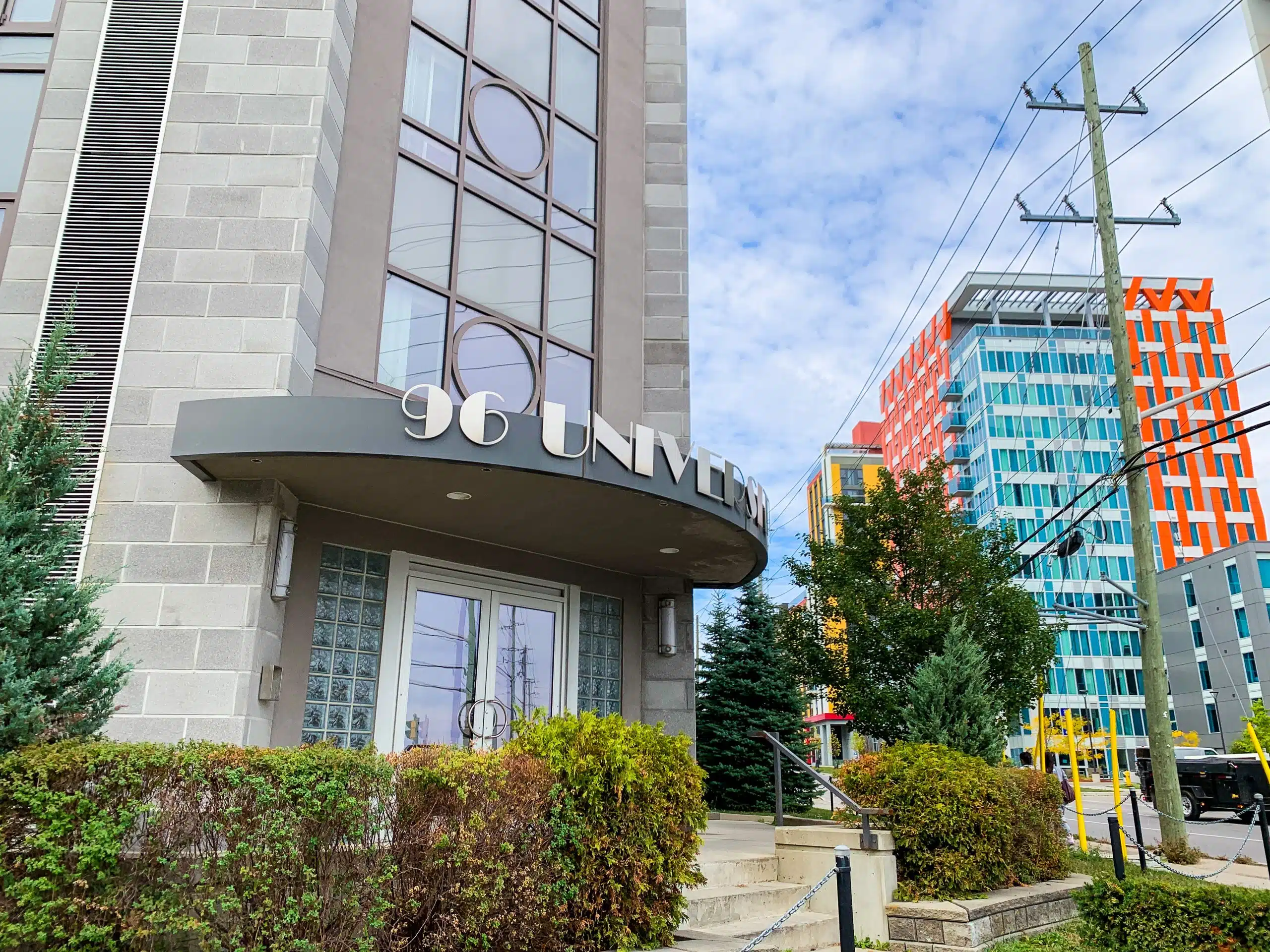 A modern building entrance with the address "96 University" displayed above the door. The entrance features glass blocks and a circular accent over the double doors. Surrounding the building are bushes and other buildings, with colorful, uniquely designed structures visible. The location is notable for hosting significant events such as the August Board Meeting.