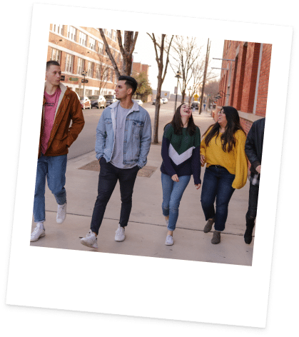 A group of five young adults walks down a city sidewalk, chatting and smiling. They wear casual clothing, and the trees and buildings in the background suggest a cool day. The photo is framed with a white Polaroid-style border and tilted slightly.