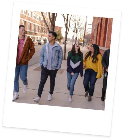 A group of five young adults walks down a city sidewalk, chatting and smiling. They wear casual clothing, and the trees and buildings in the background suggest a cool day. The photo is framed with a white Polaroid-style border and tilted slightly.