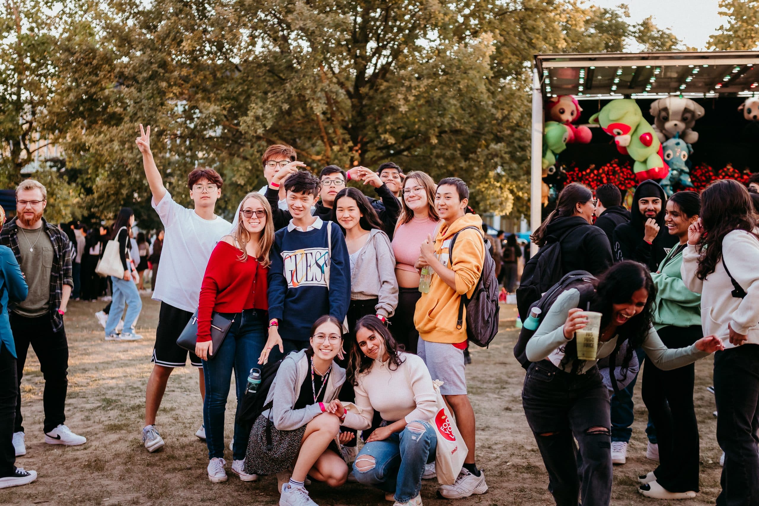 A group of smiling individuals pose for a photo at an outdoor event with trees and a game stall featuring stuffed animals in the background. Some are standing while others crouch in front, creating a lively and friendly atmosphere.