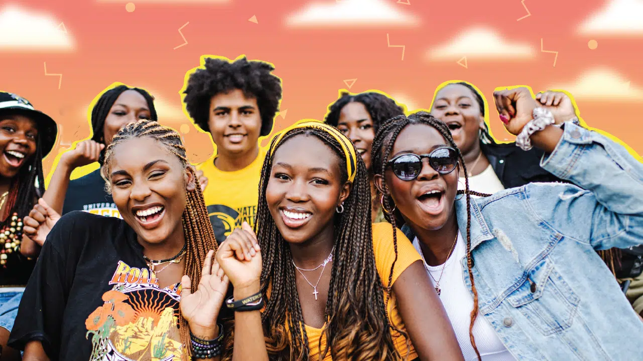 A group of smiling young adults pose energetically in front of an illustrated sunset background with stylized clouds. They display lively expressions and gestures, wearing casual, vibrant clothing. The atmosphere at the First Year Fair is joyful and celebratory, marking it as the best one ever.