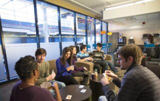 Seven people are sitting in a lounge area with large windows, engaged in a card game. Some are holding cards while others have drinks. Amidst the casual and relaxed atmosphere, a 2nd year student is giving tips near the coffee machine about how to make the most of Reading Week.