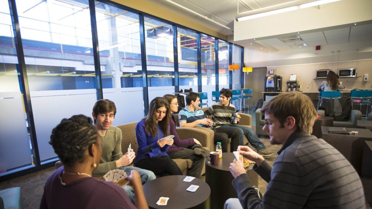 Seven people are sitting in a lounge area with large windows, engaged in a card game. Some are holding cards while others have drinks. Amidst the casual and relaxed atmosphere, a 2nd year student is giving tips near the coffee machine about how to make the most of Reading Week.