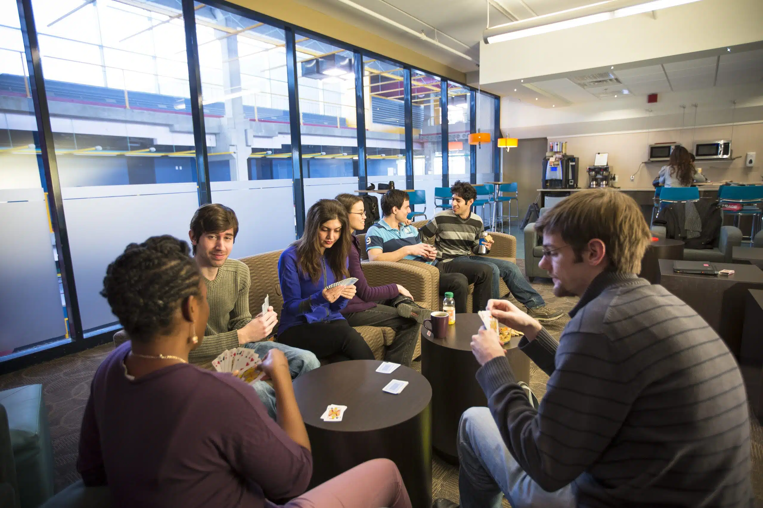 Seven people are sitting in a lounge area with large windows, engaged in a card game. Some are holding cards while others have drinks. Amidst the casual and relaxed atmosphere, a 2nd year student is giving tips near the coffee machine about how to make the most of Reading Week.