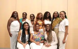 A group of ten women smiling and posing together in front of a light-colored wooden wall. Nine of them stand, while one woman in the center sits. The women, gathered for Clubs and Societies Day, are dressed in colorful and neutral outfits, some wearing traditional patterns.