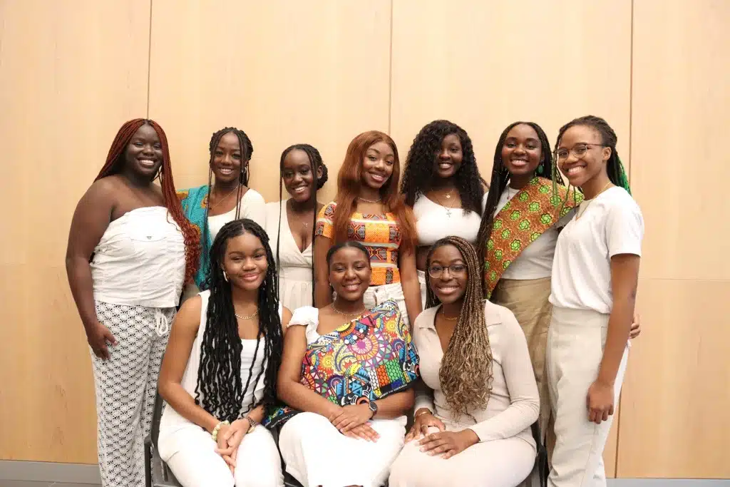 A group of ten women smiling and posing together in front of a light-colored wooden wall. Nine of them stand, while one woman in the center sits. The women, gathered for Clubs and Societies Day, are dressed in colorful and neutral outfits, some wearing traditional patterns.