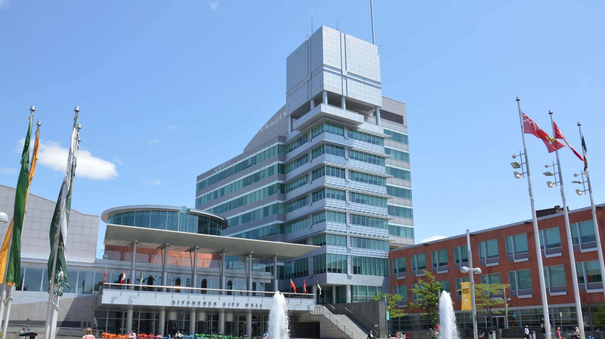 A modern, multi-story building with glass windows and a distinctive square tower stands under a clear blue sky. The building, adorned with flags and fountains, features a sign that reads "City Hall" in both English and French. This landmark in the Region of Waterloo is central to student engagement initiatives.