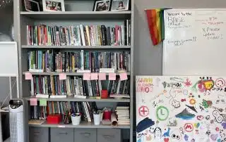 A bookshelf holds numerous books arranged neatly, some with pink sticky notes. Framed photos rest on top. To the right, a whiteboard has text and a colorful Pride flag, showing support for BIPOC talent. Below, a large canvas displays a mix of sketches, stickers, and doodles in various colors.