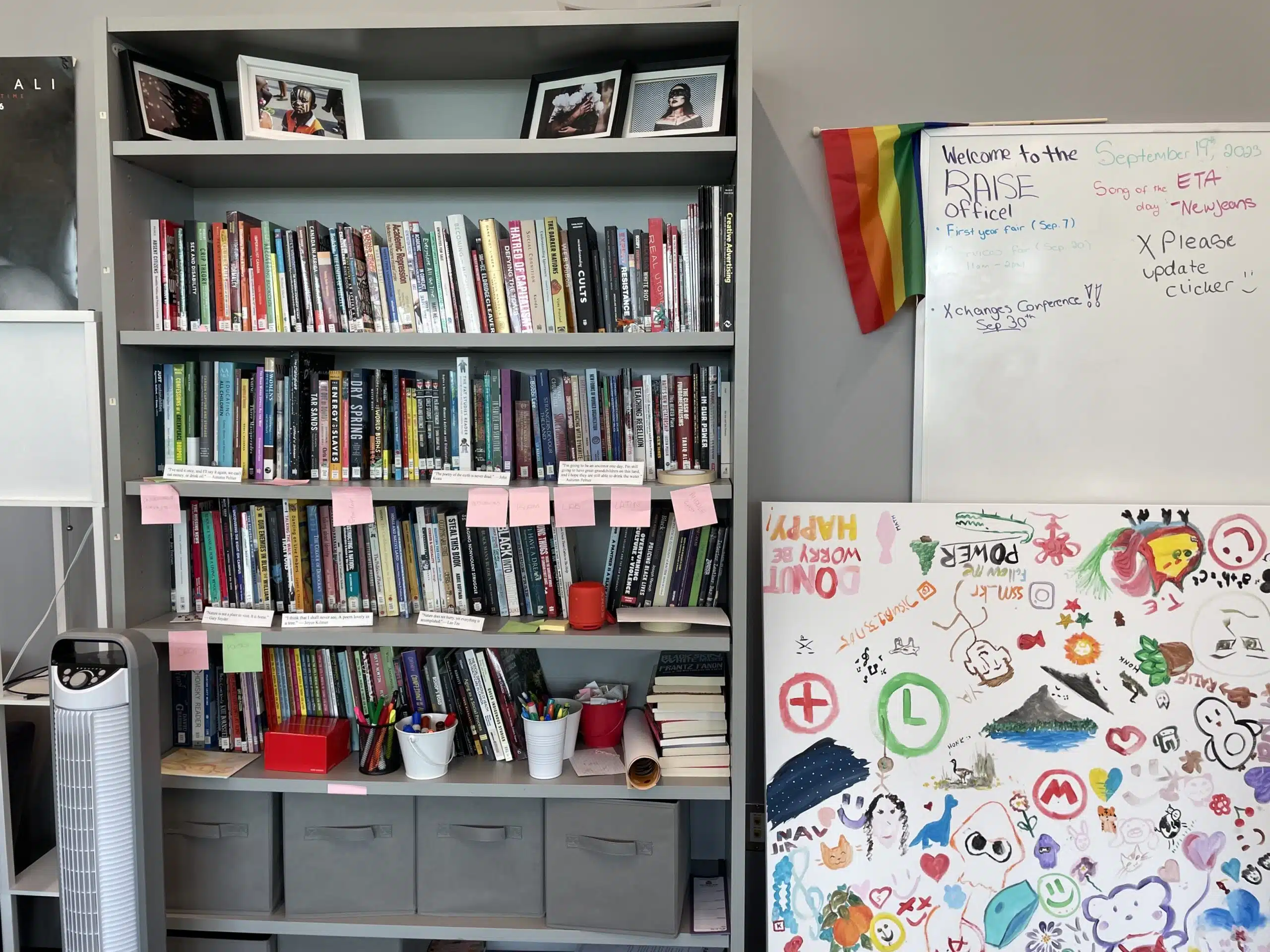 A bookshelf holds numerous books arranged neatly, some with pink sticky notes. Framed photos rest on top. To the right, a whiteboard has text and a colorful Pride flag, showing support for BIPOC talent. Below, a large canvas displays a mix of sketches, stickers, and doodles in various colors.