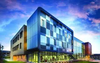 Modern, multi-story building with a striking design, featuring a mix of blue and green glass panels and large windows. The structure is part of an educational institution with the name "Université de l'Ontario français" visible on the lower part of the building. The sky is pink and blue at sunset, setting the stage for the launch of their innovative GBDA Pilot Bus Program.