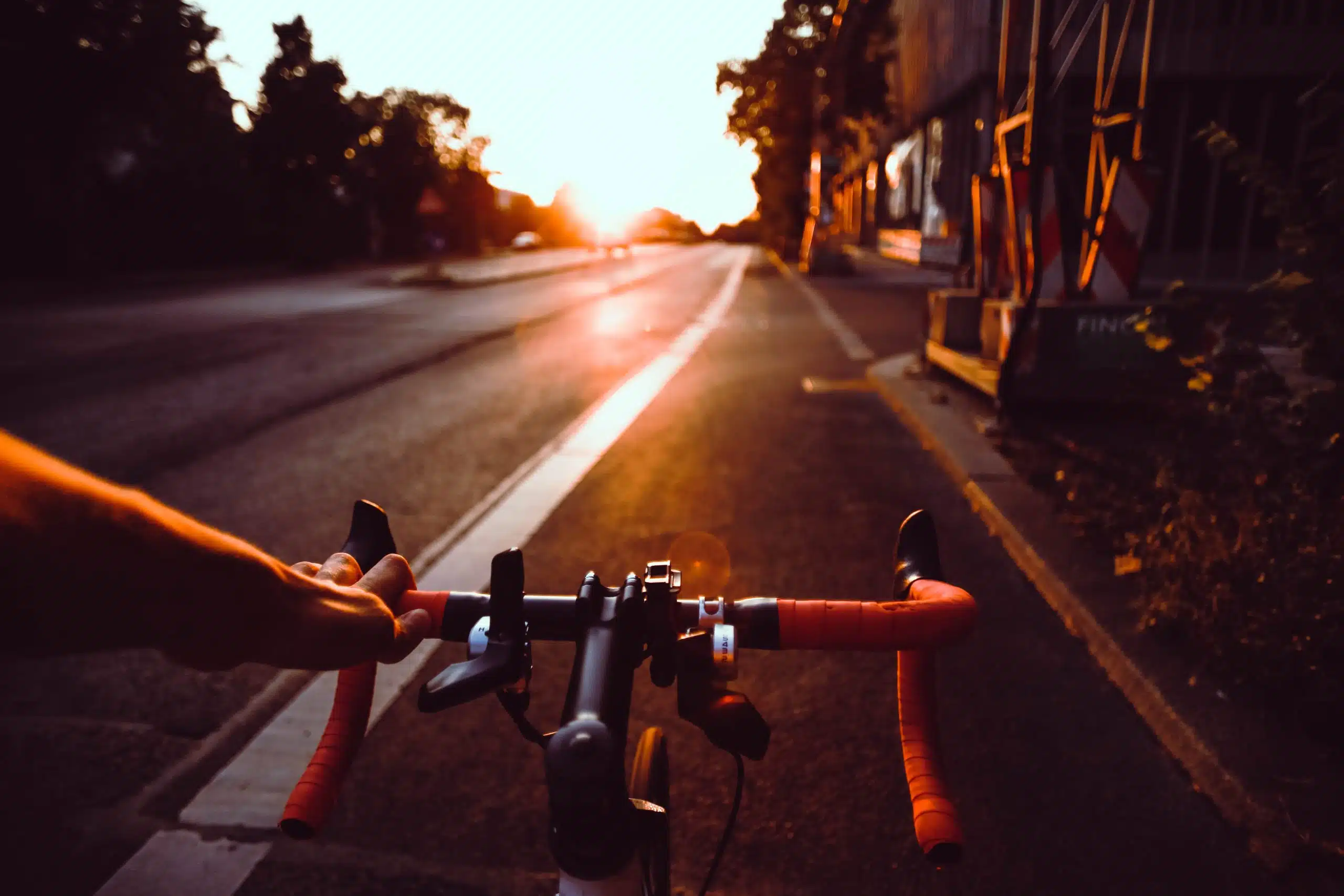 A person's hand gripping the handlebars of a bike on a road during sunset. The sun is low on the horizon, casting a warm glow on the surroundings, which includes trees and buildings on the right side. The mostly empty road enhances the serene atmosphere, with nature at its centre.