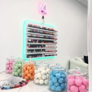 A nail salon in Uptown Waterloo features a wall display filled with various nail polish colors. Below, glass jars are filled with colorful bath bombs in green, orange, white, blue, and pink. A neon sign above the display reads "GRL PWR." A color palette sample fan is also visible on the table. Explore this vibrant spot!
